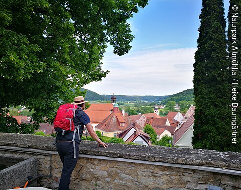 Wanderer am Wallfahrerweg in Greding Ein Wanderer mit roten Rucksack steht an einer Mauer und lässt seinen Blick über Greding schweifen. Die Stadt ist von zwei Bäumen eingerahmt.