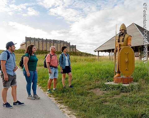 Wanderer stehen vor der Keltenstatue aus Holz bei der Freilichtanlage mit der Rekonstruktion des des ehemaligen keltischen Fürstenhofs.