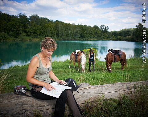Eine Reiterin studiert die Wanderreitkarte. Im Hintergund grasen an einem kleinen See zwei Pferde.