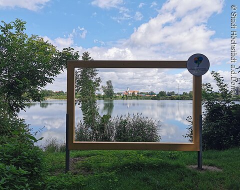 Herzogin-Anna-Rundweg Ein am Seeufer stehender, großer, hölzerner Bilderrahmen durch den man den See und die am anderen Seeufer liegende Ortschaft sieht.