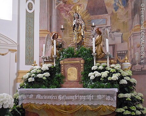Der mit weißen Blumen geschmückte Marienaltar. In der Mitte die Marienstatue mit Kind. Zu ihren Füßen knien beidseitig Engel mit Kerzen.