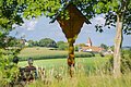 Aussicht auf Utzwingen mit seinem Kirchturm. Im Bildvordergrund sieht man ein hölzernes Wegkreuz und eine Wanderin, die sich auf einer Bank ausruht und die Aussicht genießt.
