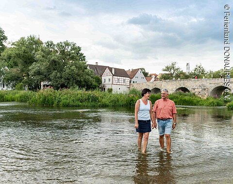 Ein Paar watet am Wörnitzstrand durchs Wasser. Im Hintergrund sieht man die steinerne Brücke und Häuser von Harburg.