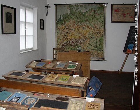 Blick in ein altes Schulzimmer mit alten, hölzernen Schulbänken und einer Landkarte an der Wand. Auf dem Tischen liegen Bücher und Schreibtafeln.