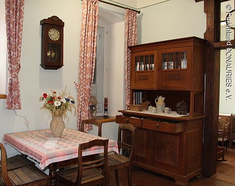 Eine Wohnstube mit großem Schrank, einem Tisch mit Stühlen und eine über dem Tisch an der Wand angebrachte hölzerne Uhr