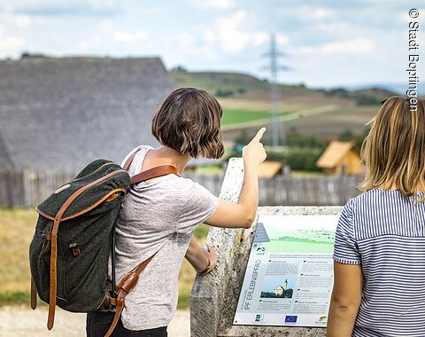 Wanderer am keltischen Freilichtmuseum unterhalb des Ipfs. Zwei Wanderinnen lesen eine Infotafel.