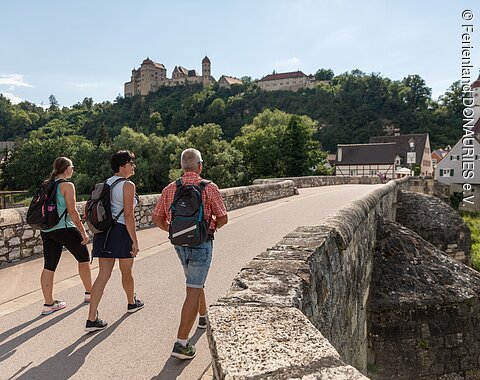 Drei Wanderer laufen über die Steinerne Brücke in Harburg. Links oben im Bild sieht man Schloss Harburg.