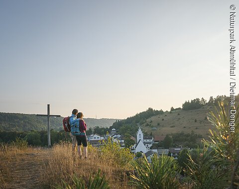 Zwei Wandernde stehen neben dem Kreuz auf dem Pfünzer Kirchberg und schauen über Pfünz zum Osterberg mit seiner Wacholderheide. Über dem Osterberg geht gerade die Sonne unter.