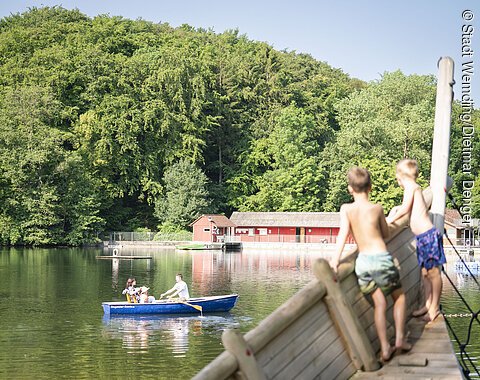 Zwei Kinder stehen auf dem Piratenschiff und beobachten ein Ruderboot, dass auf dem Waldsee in Wemding fährt.
