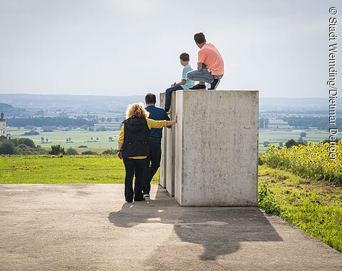 Die Kinder sitzen auf den Steinen der Zeitpyramide und die Eltern stehen daneben. Gemeinsam blicken sie in das Ries und genießen den tollen Ausblick.