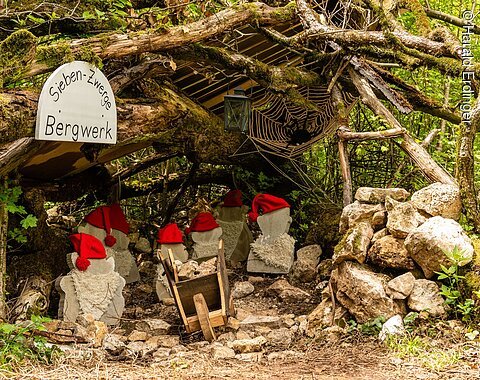 Ein Bergwerkstollen wird durch einen Gang, gebildet durch Äste dargestellt. Darin stehen Zwergenfiguren aus Stein mit roten Mützen. Rechts im Stollen sieht man eine Spinne im Netz.