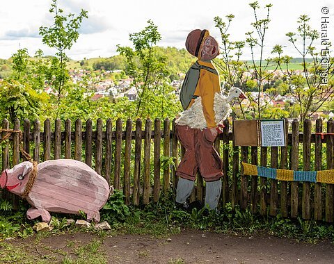 Hans im Glück, dargestellt durch eine Figur mit Gans unter dem Arm und ein Schwein aus Holz. Im Hintergrund sieht man die Stadt Harburg