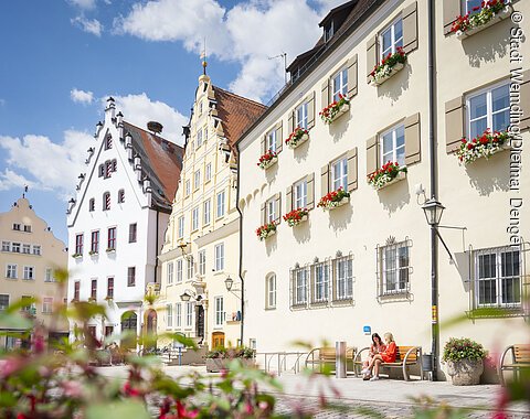 Zwei Freundinnen sitzen auf einer Bank vor dem Verwaltungsgebäude am historischen Marktplatz und unterhalten sich.