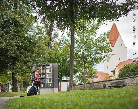 Eine Frau steht mit ihrem Hund am Bücherschrank im Stadtgraben Wemding.