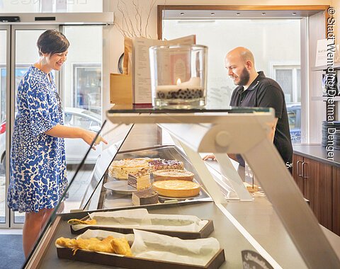 Ein Mann bedient eine Kundin an der Kuchentheke beim Lieblingsgrieche.