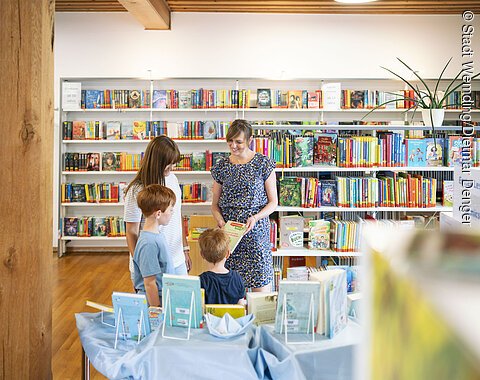 Stadtbüchereileiterin Frau Hertle berät eine Frau mit ihren Kindern zu bestimmten Büchern in der Stadtbücherei.