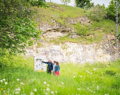 Drei Personen stehen auf einer Wiese mit hohem Gras vor einer Informationstafel. Im Hintergrund ist eine kleine Felswand zu sehen.
