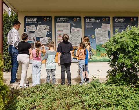 Ein Mann und eine Gruppe Kinder betrachtet Informationstafeln an einer Hauswand.