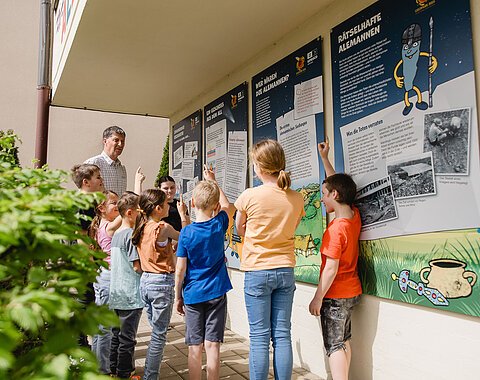 Ein Mann und eine Gruppe Kinder betrachtet Informationstafeln an einer Hauswand.