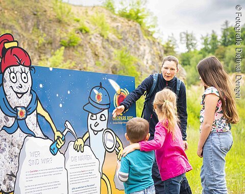 Eine Frau zeigt drei Kindern eine Informationstafel mit den Maskottchen Suevitchen und Riesitchen.