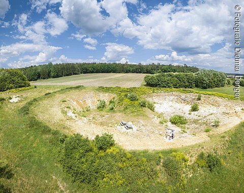 In einer flachen Grube stehen vereinzelt Büsche, während die Hänge aus bloßem Gestein bestehen. Um die Grube herum befinden sich Wälder, Äcker und Wiesen.