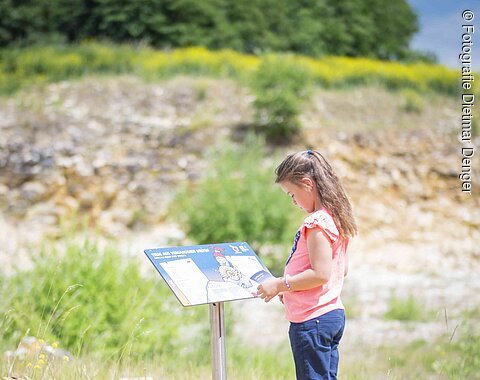 In einer flachen Grube aus Lockergestein mit vereinzelter Vegetation betrachtet ein Mädchen eine Informationstafel für Kinder.