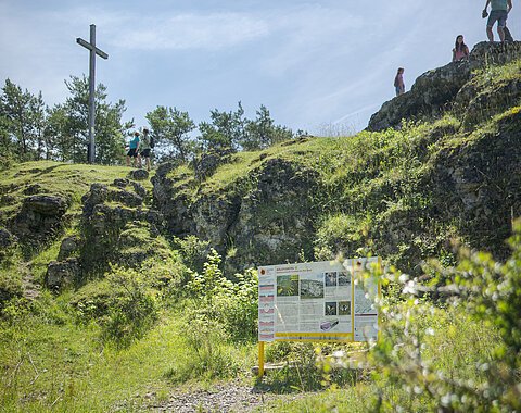 Geopark-Shooting 2021 Auf einer mit Gras bewachsenen Felsformation stehen ein Kreuz und einige Personen. Davor steht eine Informationstafel.