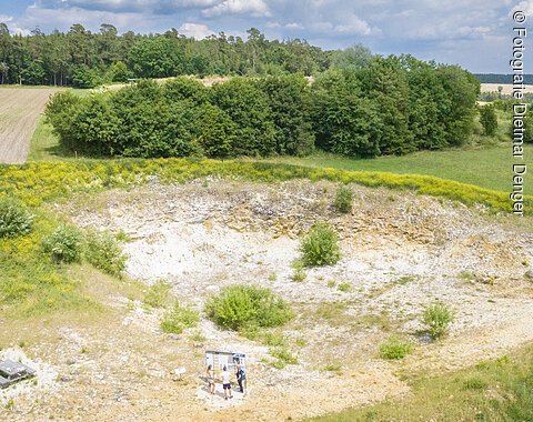In einer Grube mit vereinzelten Büschen und bloßem Gestein an den Hängen steht eine Sitzbank und eine Informationstafel.
Oberhalb der Grube befinden sich Äcker und Wald.