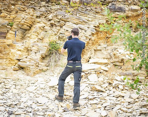 Ein Mann fotografiert mit einer Kamera eine Felswand aus plattigem Kalkgestein. Der Boden ist ebenfalls mit Bruchstücken von Kalkgesteinsplatten bedeckt.