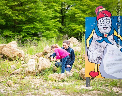 Neben einer Informationstafel mit der Figur Suevitchen klopfen ein Mädchen und eine Frau Steine.