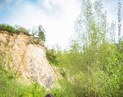 Zwei Mächen sitzen mit einem Hund auf einem Holzsteg über einen kleinen Teich. Im Hintergrund ist eine steil aufragende Felswand erkennbar.