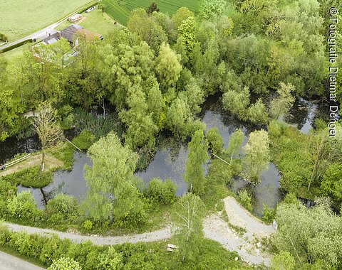 Geopark Ries Mai 2023 Das Schrägluftbild zeigt einen von Bäumen umgebenen Weiher und angrenzende Schotterwege.