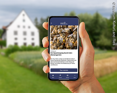 Es ist die App auf einem Handybildschirm zu sehen, der von einer Hand gehalten wird. Im Hintergrund sieht man das Museumsfeld und ein Museumsgebäude.
