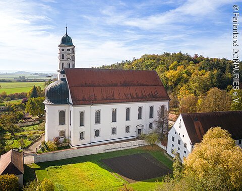 Eine große Kirche aus der Luft. Darum herum andere Gebäude, grüne Wiese und ein Hügel mit grünen Bäumen.