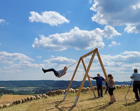 Eine Menschengruppe versammelt sich um eine schaukelnde Person auf dem Blasienberg umgeben von Schafen