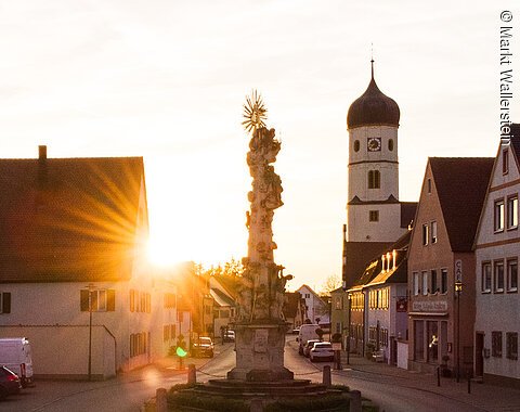 Eine barocke Stele umgeben von Häusern bei Sonnenuntergang