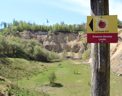 An einem Pfosten ist ein Wegweiserschild des Geopark Ries Erlebnis-Geotop Lindle angebracht. Im Hintergrund ist ein ehemaliger Steinbruch mit vielen Büschen und Bäumen zu sehen.