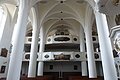 Innenansicht der Stadtpfarrkirche St. Walburga in Monheim, mit hohen weißen Säulen, gewölbter Decke, Emporen mit Verzierungen und Holzbänken.