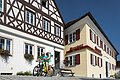 Tourist-Information der Stadt Monheim/Monheimer Alb: ein Fachwerkhaus mit drei Stockwerken, weißen Wänden und braunen Balken. Rechts davon ein gelbes zweistöckiges Gebäude mit roten Fensterläden. Zwei Personen mit Fahrrädern stehen davor und schauen eine Broschüre an.
