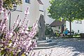 Steinfigur der heiligen Walburga auf einem gepflasterten Platz vor der Stadtpfarrkirche St. Walburga in Monheim, daneben zwei sitzende Personen auf einer Bank, dahinter ein Baum. Im Vordergrund unscharf Blumen.