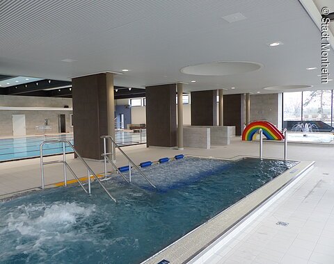 Innenansicht eines Schwimmbads mit mehreren Becken, Rutsche in Regenbogenfarben und Wasserspielen.