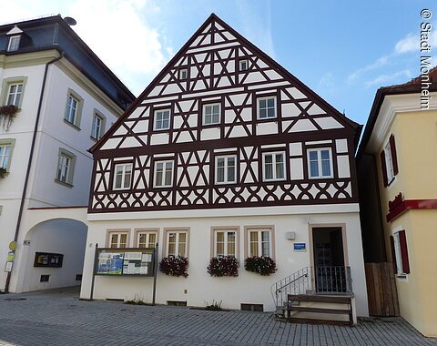 Tourist-Information der Stadt Monheim/Monheimer Alb: ein Fachwerkhaus mit drei Stockwerken, weißen Wänden und braunen Balken. Davor steht eine Sitzbank und ein Schaukasten auf einem gepflasterten Platz. Links davon ein weißes dreistöckiges Gebäude. Rechts davon ein gelbes zweistöckiges Gebäude mit roten Fensterläden.