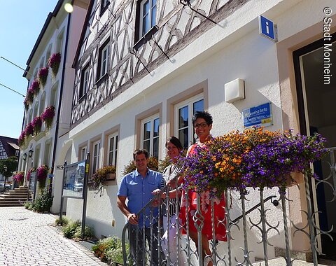 Drei Personen stehen auf einer Treppe vor einem Gebäude, der Touristinformation Monheim. Das Gebäude mit Fachwerkfassade und Blumenkästen steht auf einem gepflasterten Platz, daneben ein blaues Schild mit der Aufschrift "Tourist-Info". Im Hintergrund ein weiteren dreistöckiges Gebäude mit Blumenkästen.