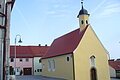 Gelbe Kapelle, die Peterskapelle in Monheim mit rotem Dach und einem Turm, umgeben von Wohnhäusern mit roten Dächern. Im Hintergrund blauer Himmel.