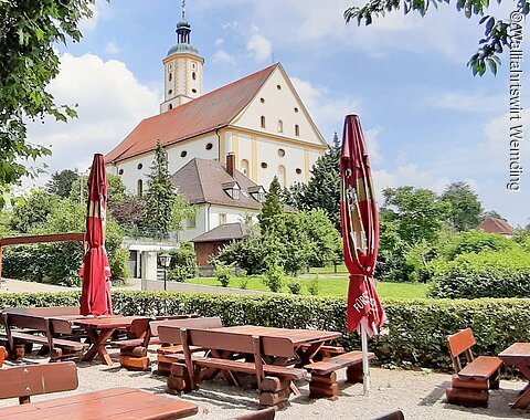 Im Vordergrund sieht man einen Teil des Biergartens mit seinen Tischen und Bänken. Im Hintergrund sieht man Wallfahrtsbasilika Maria Brünnlein.