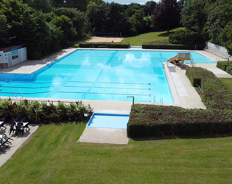 Außenansicht eines leeren Schwimmbeckens mit Sprungturm, Rutsche, Sitzbereich und umgebender Grünfläche.