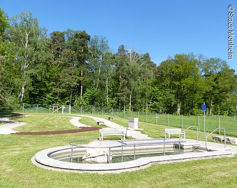 Rundes Kneippbecken in Monheim mit einem Geländer und einem Barfußpfad auf einer Wiese, umgeben von einer großen Grünfläche und Bäumen und Sitzmöglichkeiten. Ein Zaun im Hintergrund.