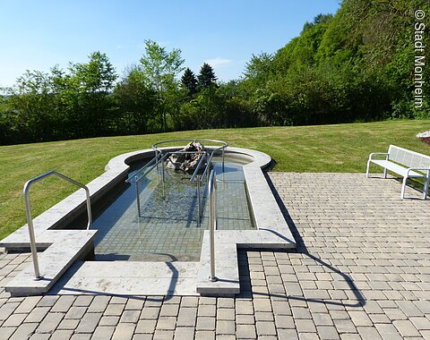 Ein befülltes Kneippbecken mit Handlauf, umgeben von Rasen und Bäumen, daneben eine weiße Bank auf Pflastersteinen.