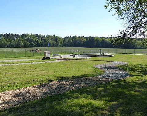 Grüne Wiese mit einem geschwungenem Barfußpfad mit Holzschnitzeln und Steinen und umzäuntem Bereich im Hintergrund, umgeben von Bäumen.