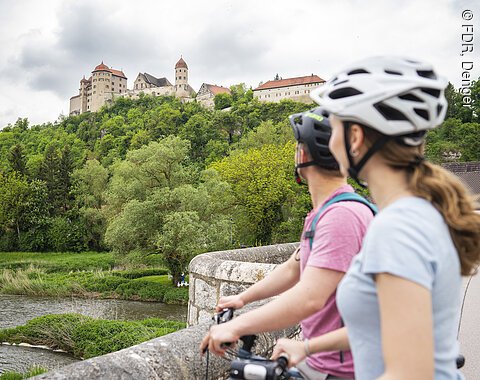 Radler auf der Steinernen Brücke in Harburg Zwei Personen mit Fahrrädern und Helmen stehen auf einer Brücke und blicken auf eine Burg auf einem Hügel.